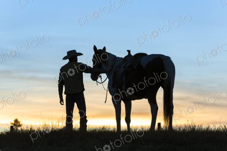 Cowboy standing next to his horse in a Prairie landscape at sunset.