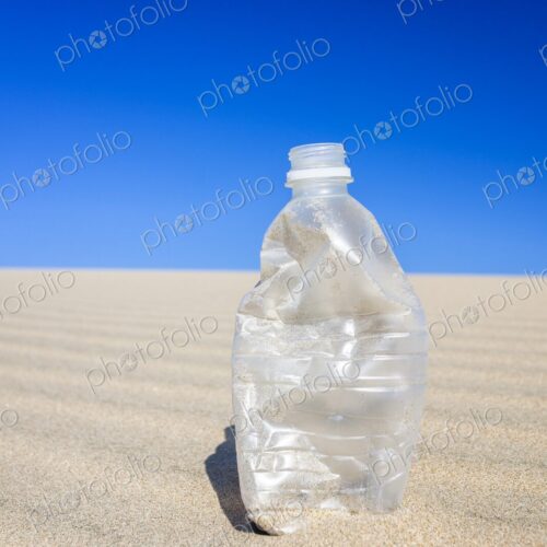 Crushed, empty plastic water bottle standing on rippled desert sand under a hot blue…