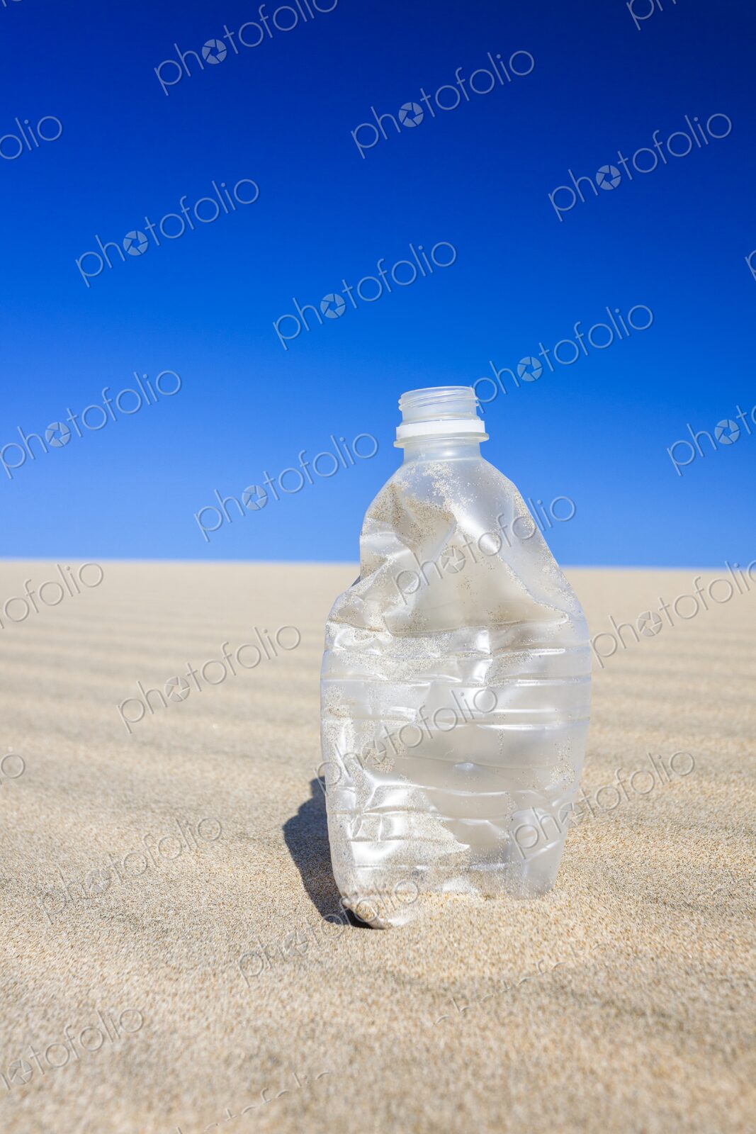 Crushed, empty plastic water bottle standing on rippled desert sand under a hot blue…