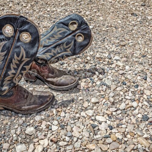 Dusty, worn leather cowboy boots rest on a rugged gravel surface, evoking Western…