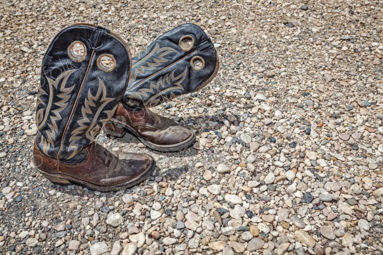 Dusty, worn leather cowboy boots rest on a rugged gravel surface, evoking Western…