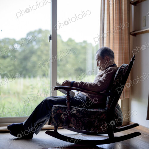 Elderly man sitting in rocking chair by a window, reading book.