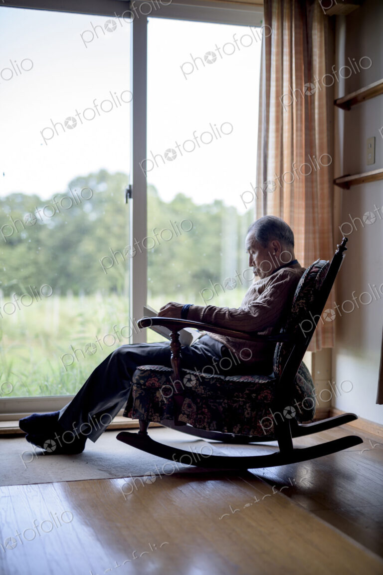 Elderly man sitting in rocking chair by a window, reading book.