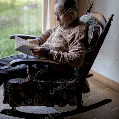 Elderly man sitting in rocking chair by a window, reading book.