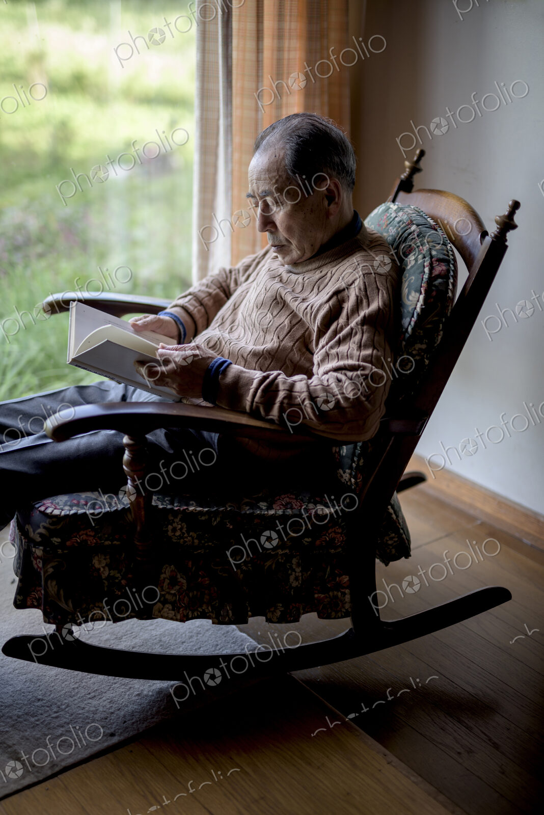Elderly man sitting in rocking chair by a window, reading book.