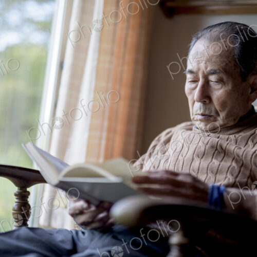 Elderly man sitting in rocking chair by a window, reading book.