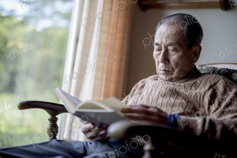 Elderly man sitting in rocking chair by a window, reading book.