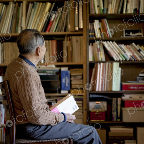 Elderly man sitting on chair in front of bookcase, holding book.