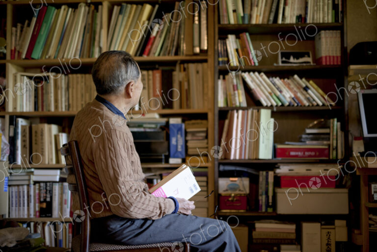 Elderly man sitting on chair in front of bookcase, holding book.