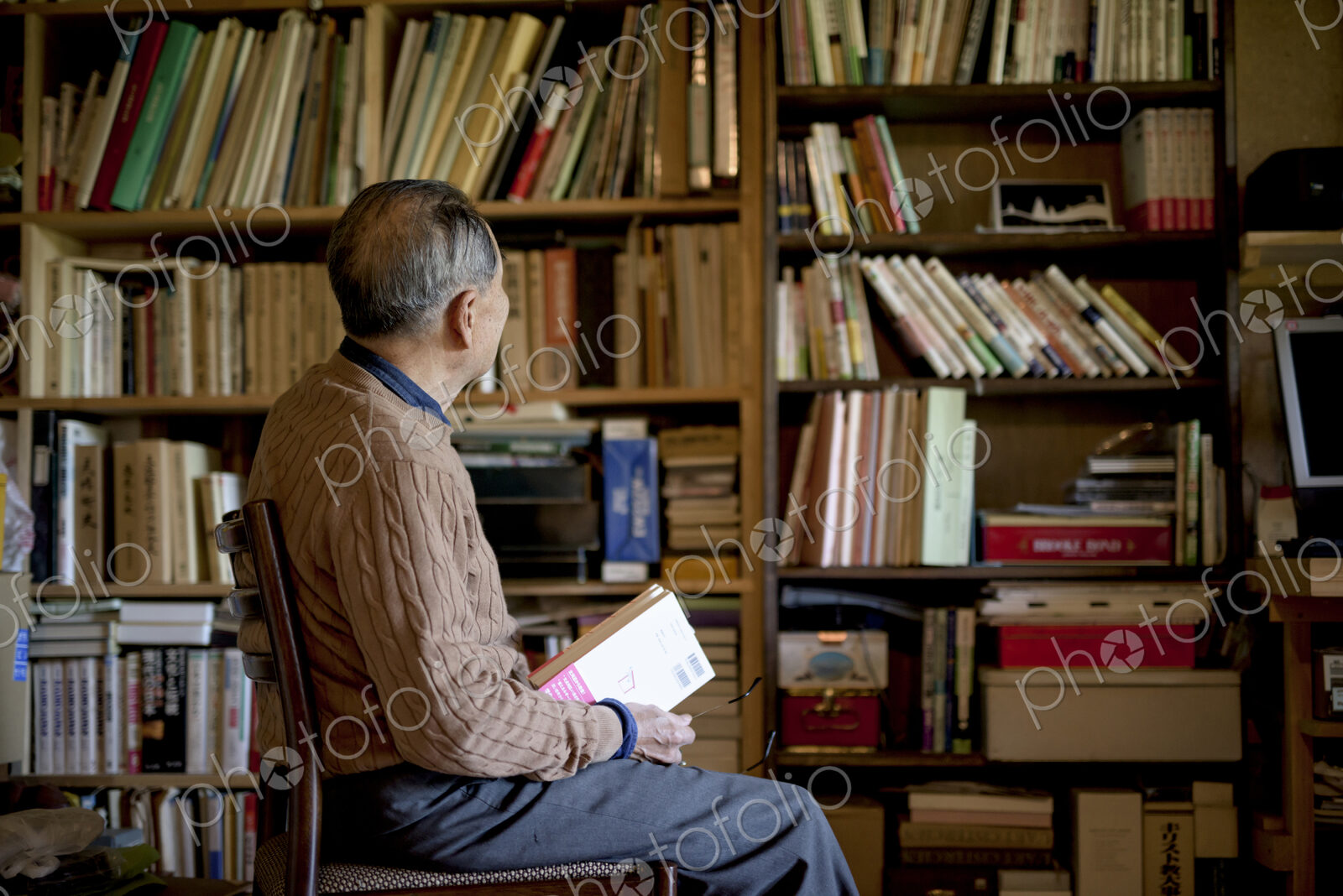 Elderly man sitting on chair in front of bookcase, holding book.