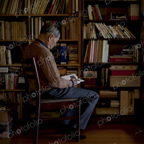 Elderly man sitting on chair in front of bookcase, reading book.