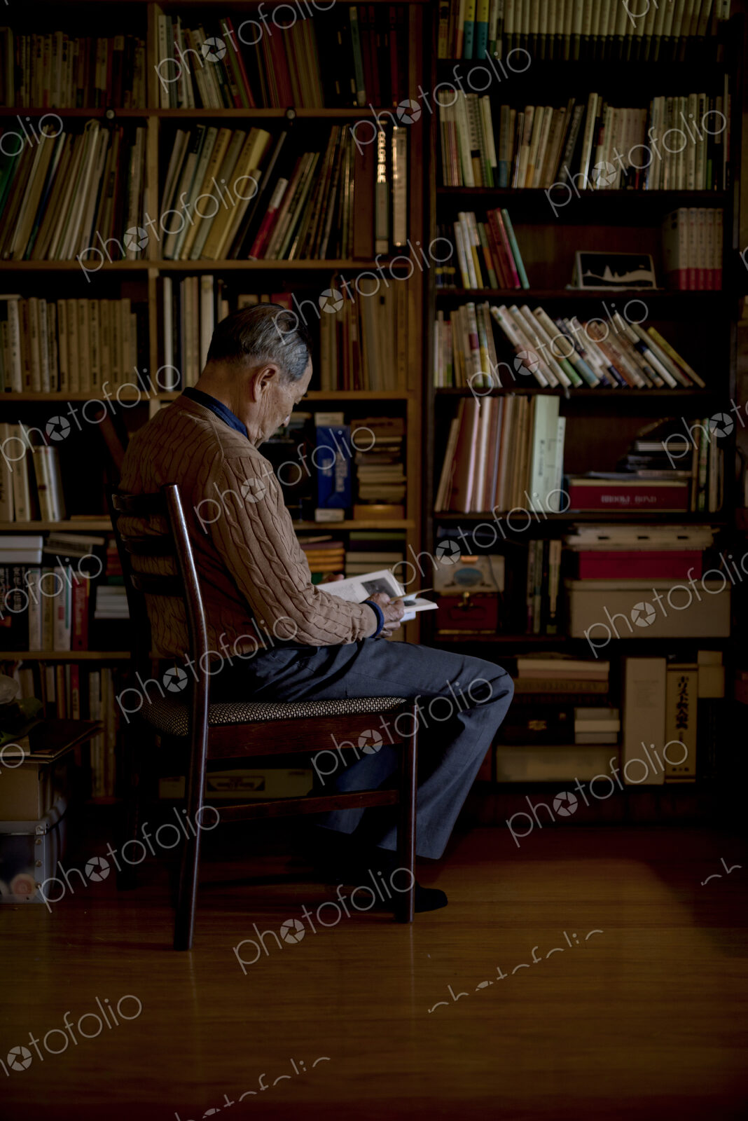 Elderly man sitting on chair in front of bookcase, reading book.