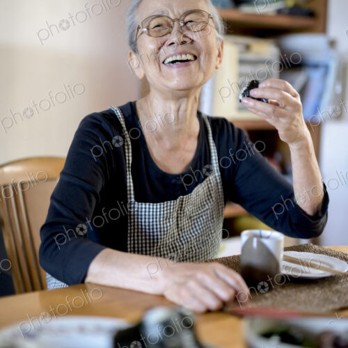 Elderly woman sitting at kitchen table, eating sushi, smiling at camera. Elderly woman sitting at kitchen table, eating sushi, smiling at camera.