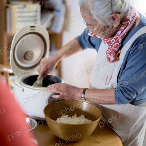 Elderly woman standing at a table in a kitchen, spooning sushi rice from rice cooker… Elderly woman standing at a table in a kitchen, spooning sushi rice from rice cooker…