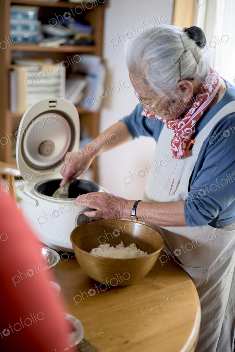 Elderly woman standing at a table in a kitchen, spooning sushi rice from rice cooker…