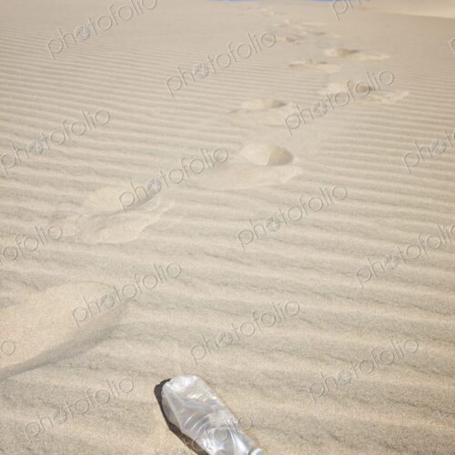 Empty plastic water bottle on rippled sand dune with footprints trailing into the…