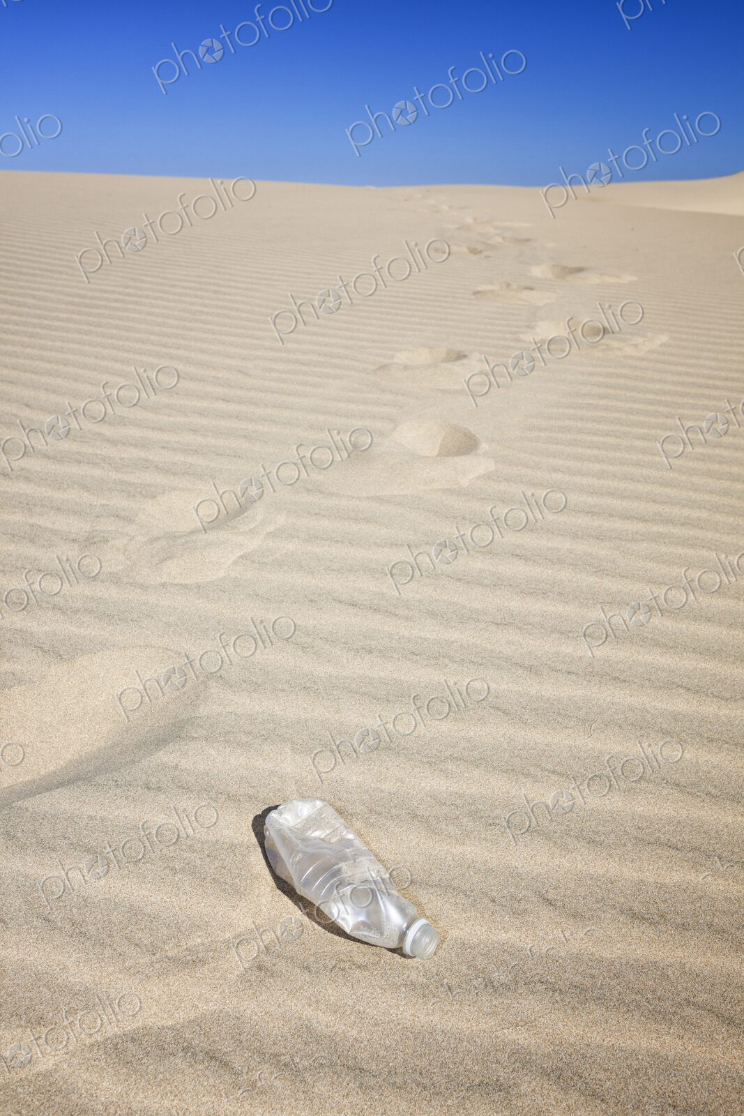 Empty plastic water bottle on rippled sand dune with footprints trailing into the…