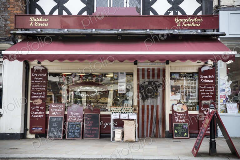 Exterior view of a butcher shop with red awning, blackboards on pavement advertising…