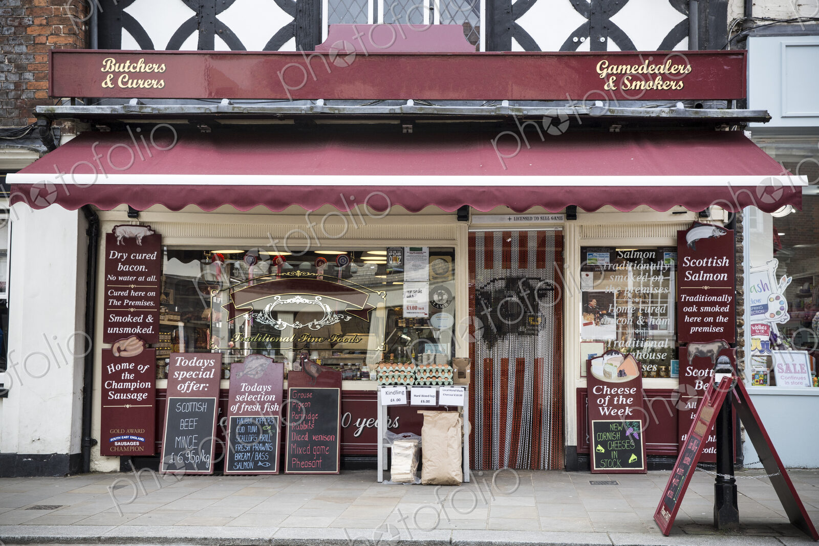 Exterior view of a butcher shop with red awning, blackboards on pavement advertising…