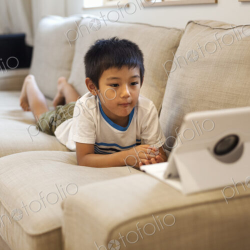 Family home. A boy lying on a sofa watching a digital tablet.