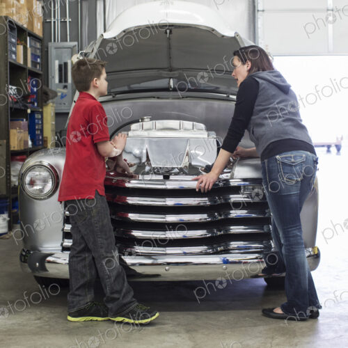 Female car mechanic talks to a boy about a car engine in a classic car repair shop.