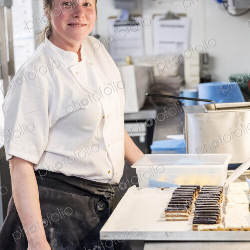 Female chef wearing chef's whites and apron standing at counter with tray of cakes in… Female chef wearing chef's whites and apron standing at counter with tray of cakes in…