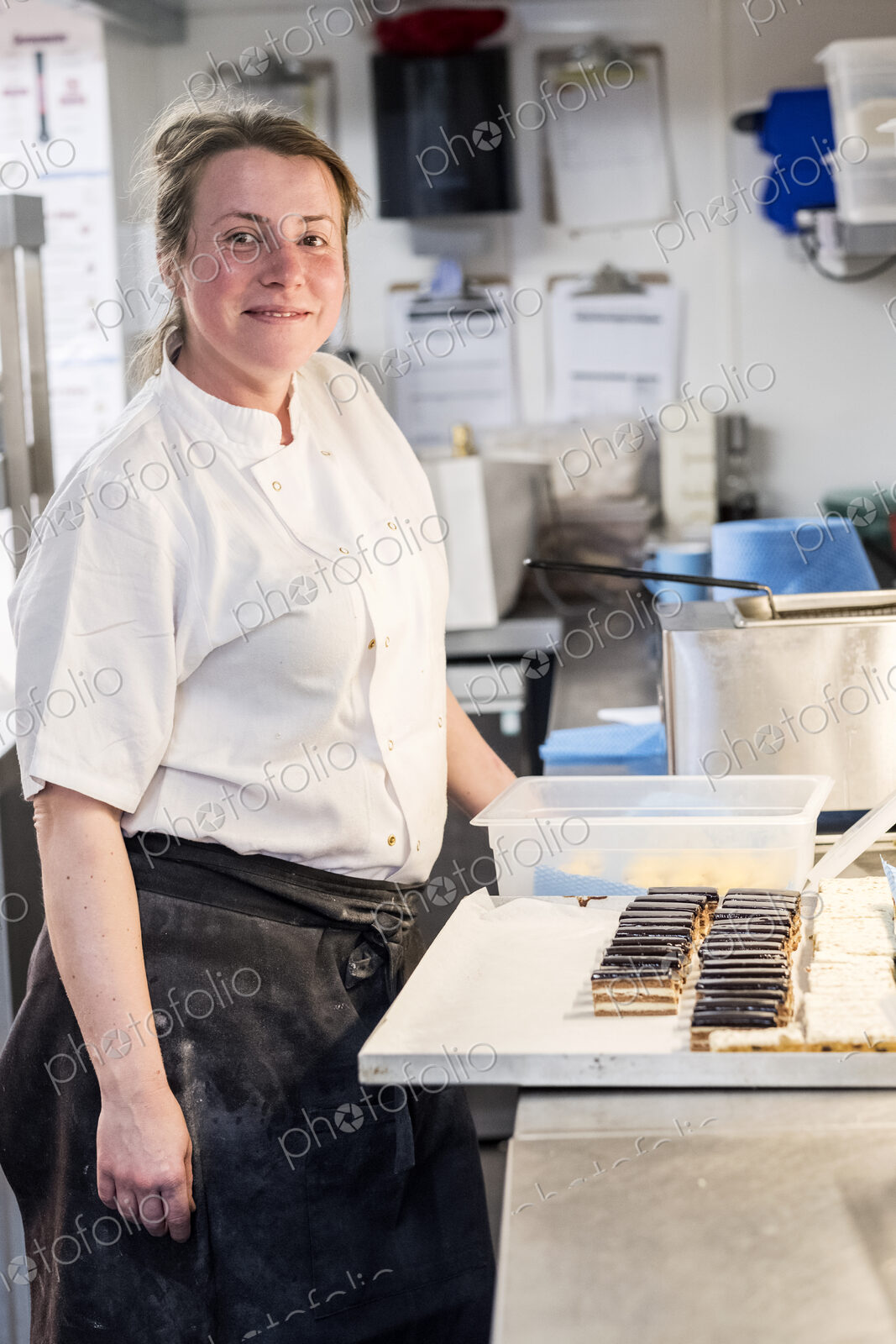 Female chef wearing chef’s whites and apron standing at counter with tray of cakes in…
