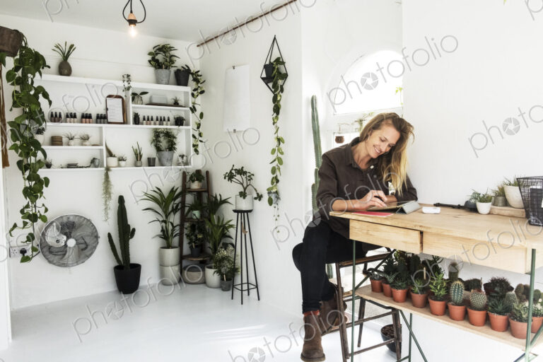 Female owner of plant shop sitting at table, working on digital tablet, a selection of…