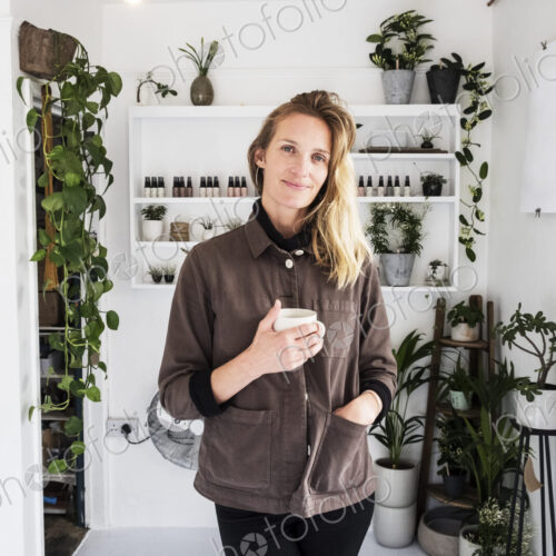 Female owner of plant shop smiling at camera, a selection of plants on wooden shelves. Female owner of plant shop smiling at camera, a selection of plants on wooden shelves.