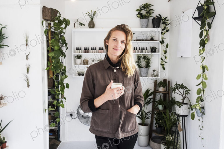 Female owner of plant shop smiling at camera, a selection of plants on wooden shelves.