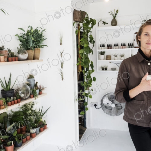 Female owner of plant shop smiling at camera, a selection of plants on wooden shelves. Female owner of plant shop smiling at camera, a selection of plants on wooden shelves.