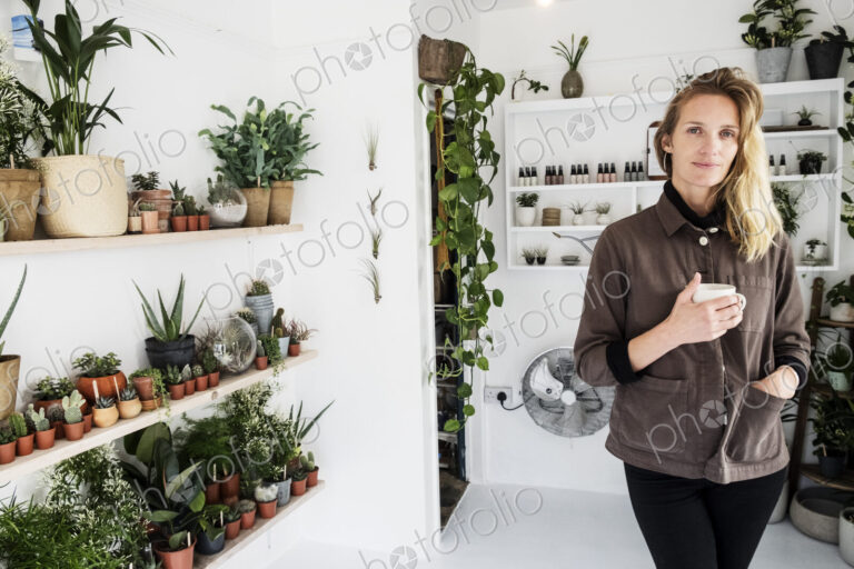 Female owner of plant shop smiling at camera, a selection of plants on wooden shelves.