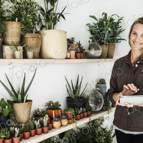 Female owner of plant shop standing next to a selection of plants on wooden shelves,… Female owner of plant shop standing next to a selection of plants on wooden shelves,…