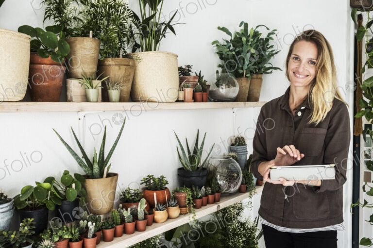 Female owner of plant shop standing next to a selection of plants on wooden shelves,…
