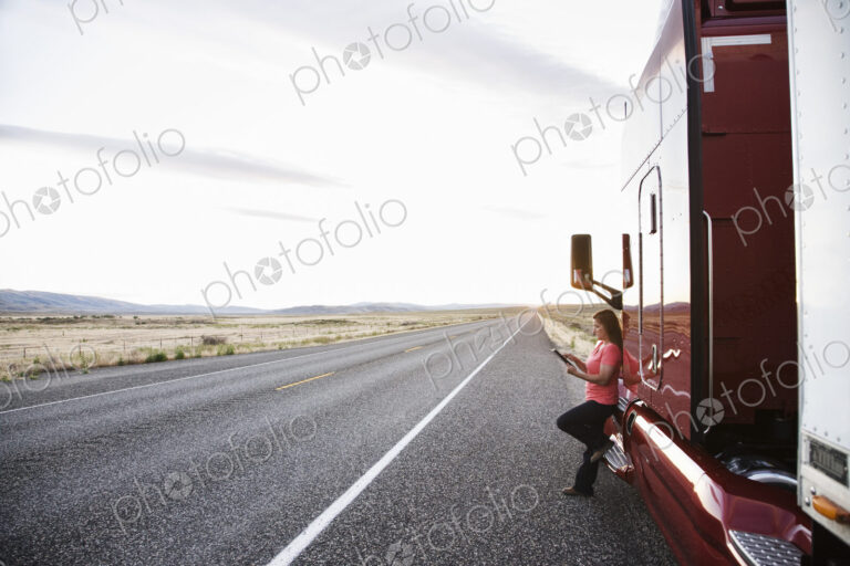 Female truck driver standing next to the cab of her truck near the highway in eastern…