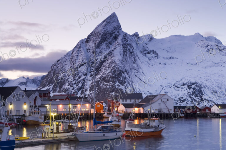 Fishing boats moored in harbour of settlement, with snow-capped mountains behind.