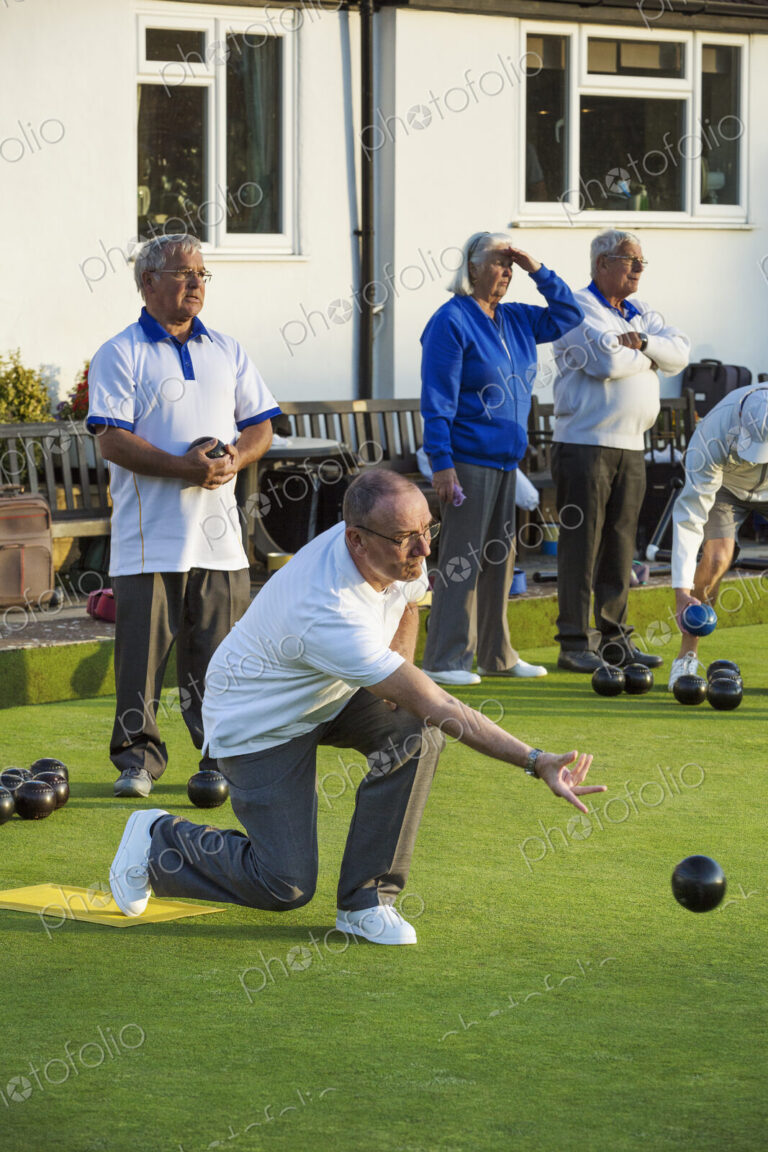 Four people at a lawn bowls match or game. One man kneeling and delivering a bowls…