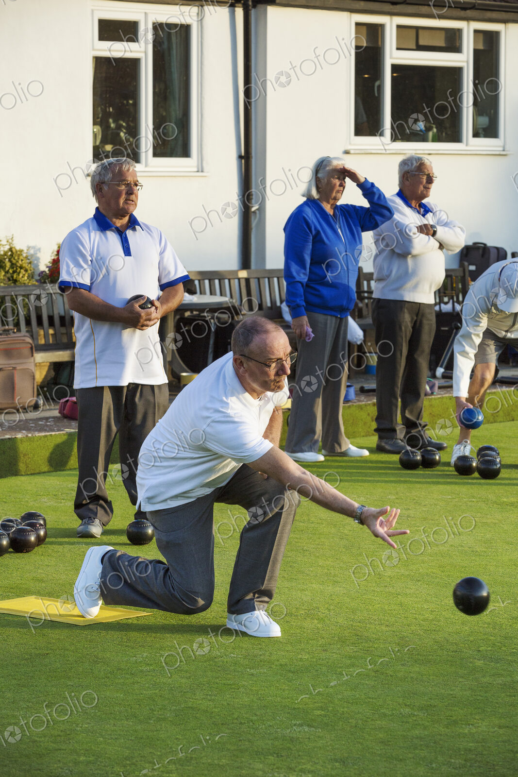 Four people at a lawn bowls match or game. One man kneeling and delivering a bowls…