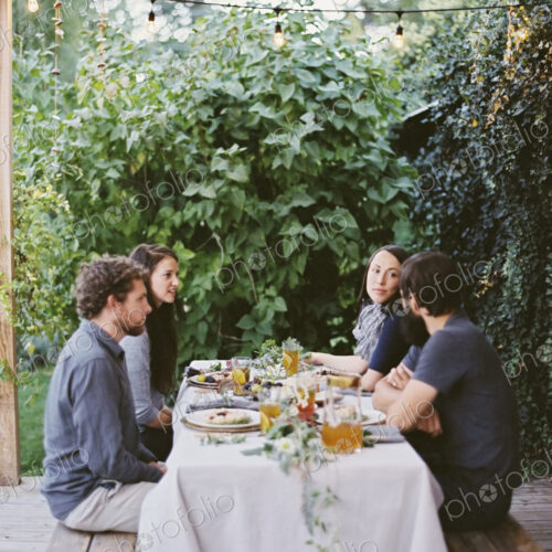 Four people seated at a table in the garden. Place settings and decorations on a white… Four people seated at a table in the garden. Place settings and decorations on a white…
