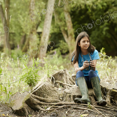 Girl birdwatching. Sitting on a tree stump holding binoculars.
