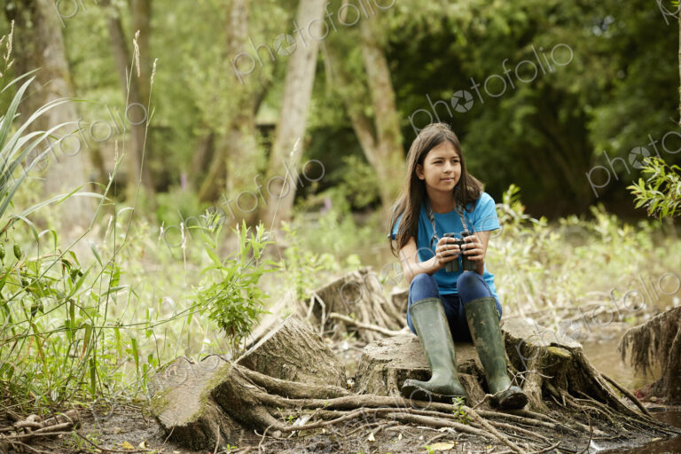 Girl birdwatching. Sitting on a tree stump holding binoculars.