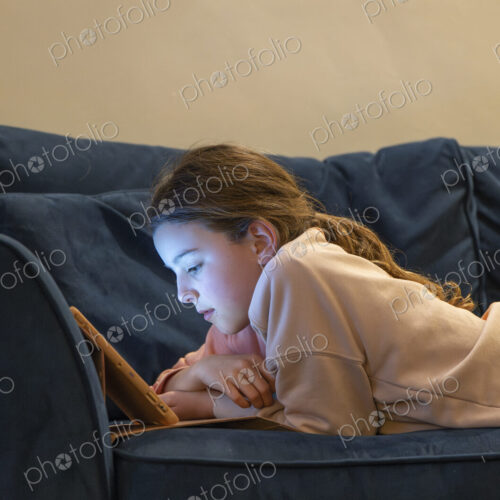 Girl relaxing on sofa with face lit up by screen of tablet.