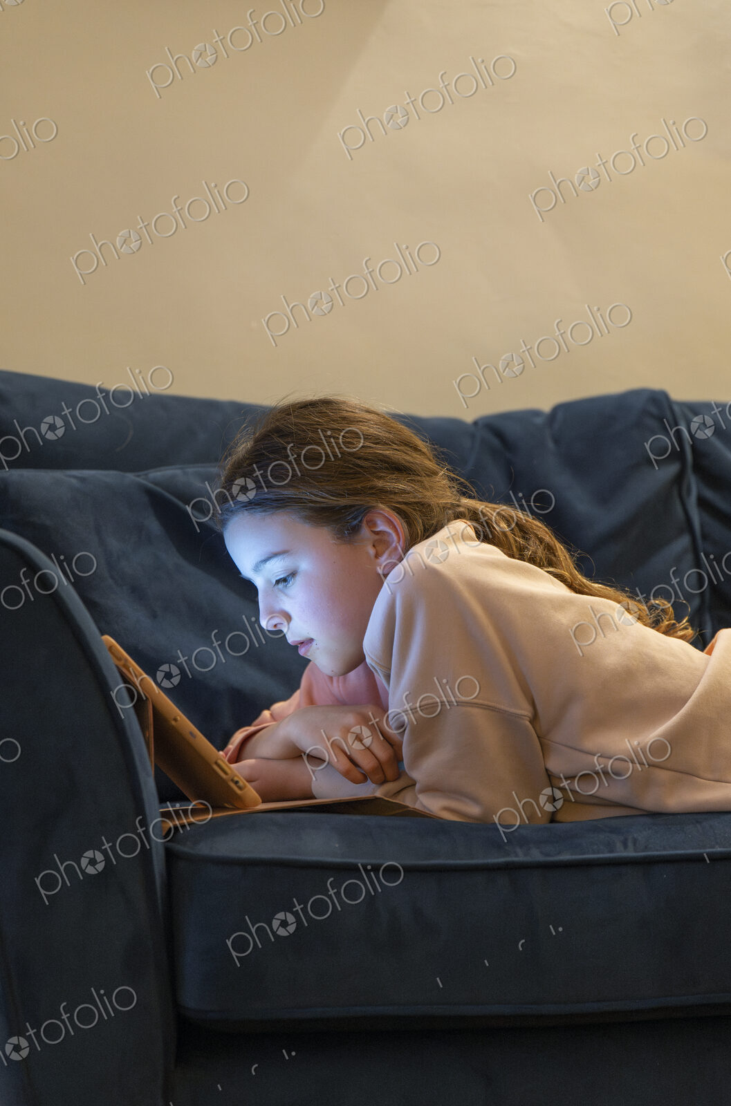 Girl relaxing on sofa with face lit up by screen of tablet.