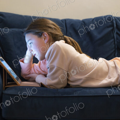 Girl relaxing on sofa with face lit up by screen of tablet.