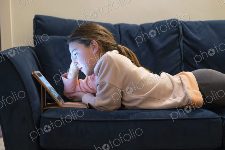 Girl relaxing on sofa with face lit up by screen of tablet.