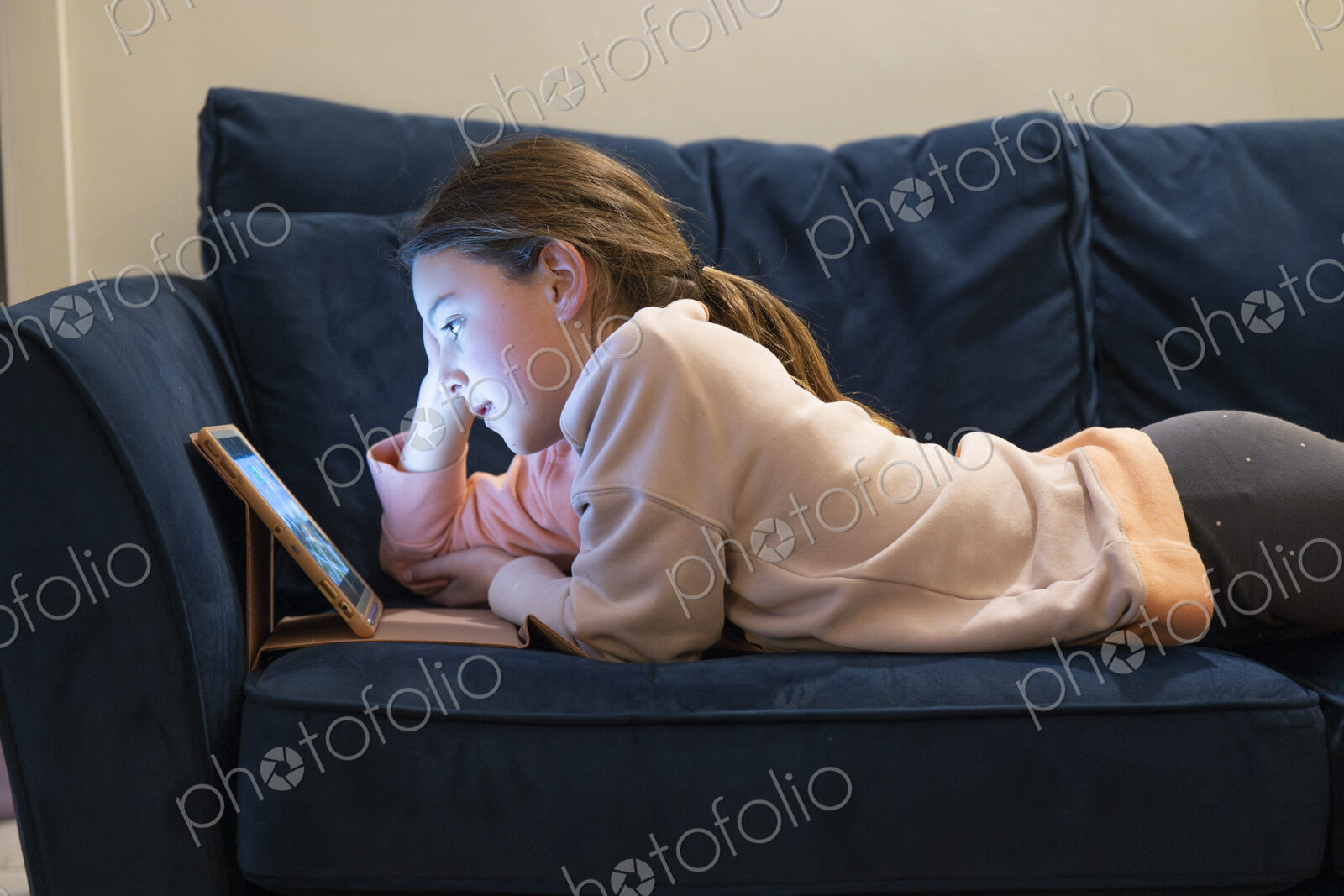 Girl relaxing on sofa with face lit up by screen of tablet.