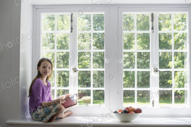 Girl sitting by a window, reading a book.