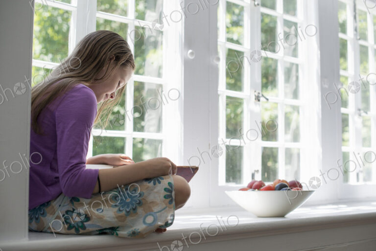 Girl sitting by a window, reading a book.