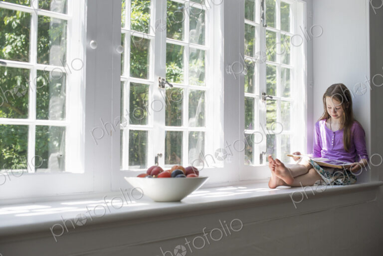Girl sitting by a window, reading a book.