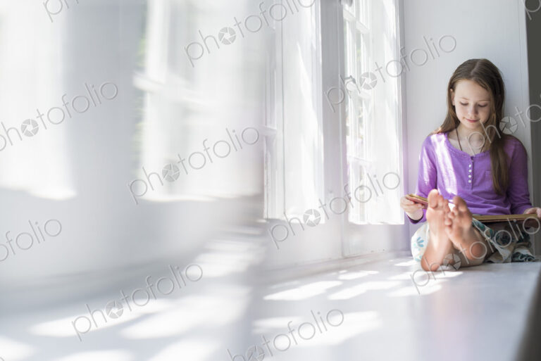 Girl sitting by a window, reading a book.